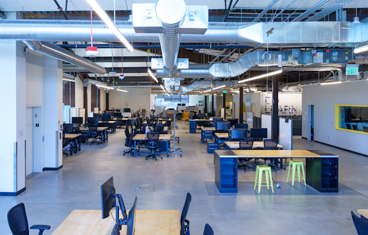 A spacious, modern office with high ceilings and exposed ductwork. The room features multiple desks with computers, ergonomic chairs, and a few green stools. The lighting is bright, and the floor is polished concrete.