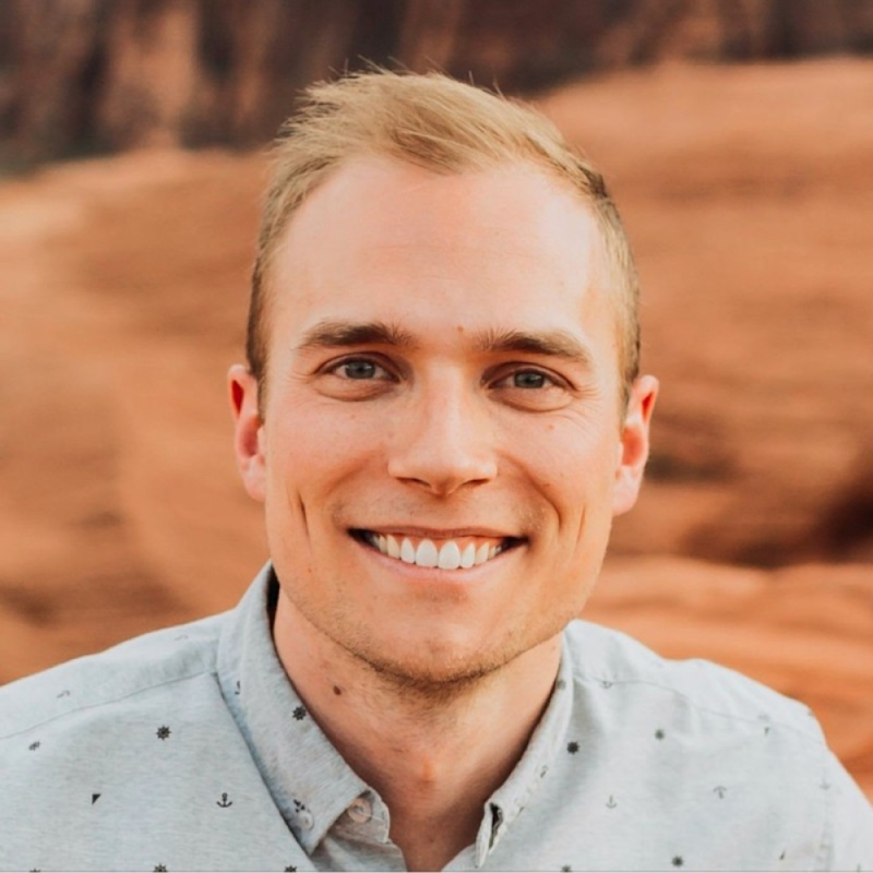 Smiling person with short blonde hair wearing a light-colored shirt, standing outdoors with a blurred natural background.