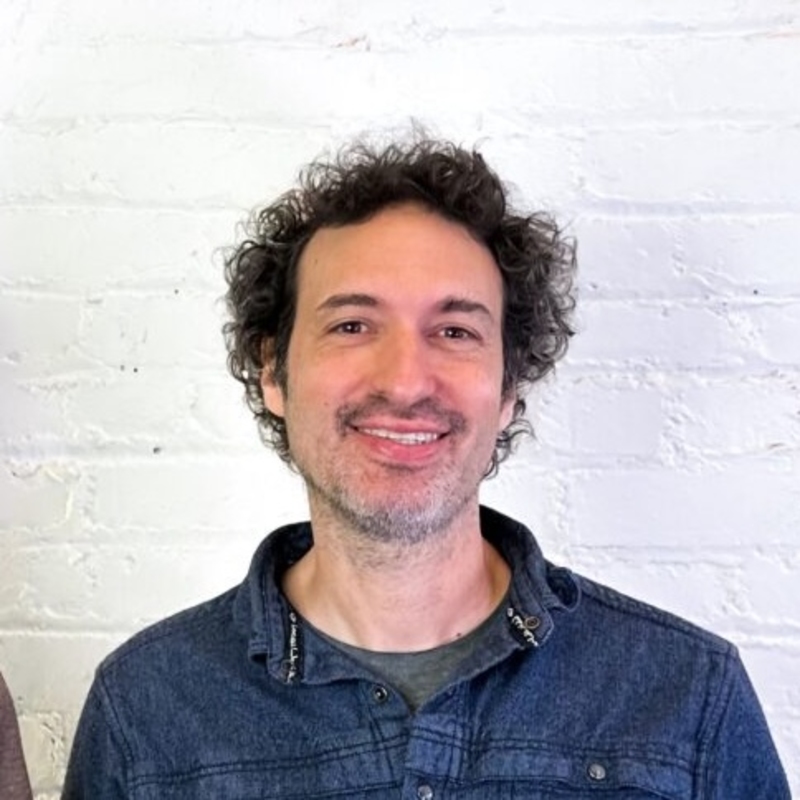 A person with curly hair and a beard smiles at the camera. They are wearing a denim shirt over a grey t-shirt, standing in front of a white brick wall.