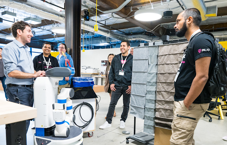 A group of people in a modern office space engage in conversation. Some wear lanyards and casual attire. In the foreground, a robot has people’s attention, suggesting a tech-related discussion or presentation.