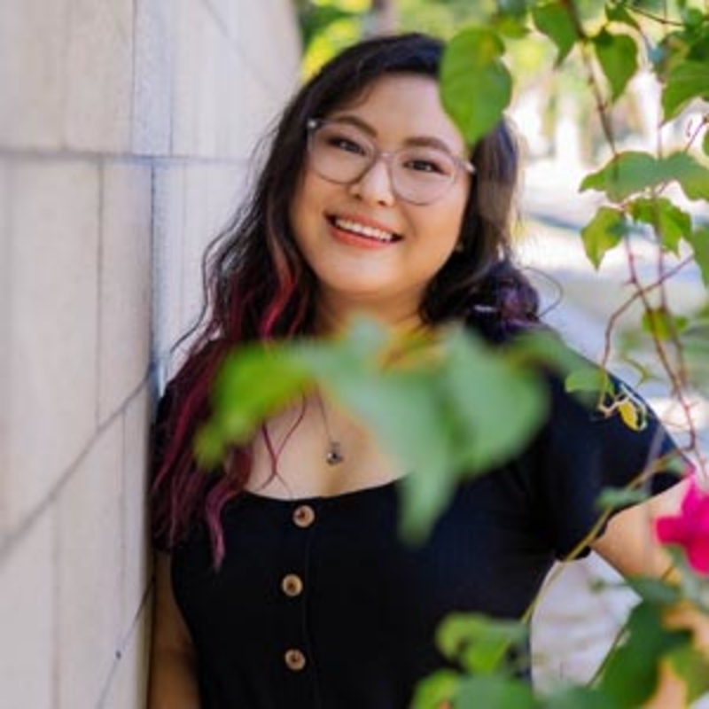 A person with long, wavy hair, wearing glasses and a black shirt, smiles while leaning against a sunlit wall. Green leaves and pink flowers are in the foreground, adding a touch of nature to the scene.