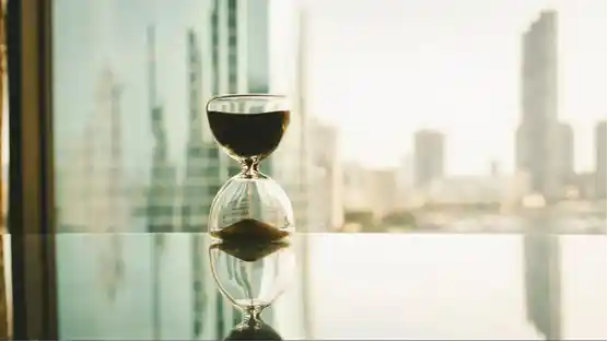 An hourglass with dark sand sits on a reflective glass surface in front of large city windows, with tall buildings blurred in the background.