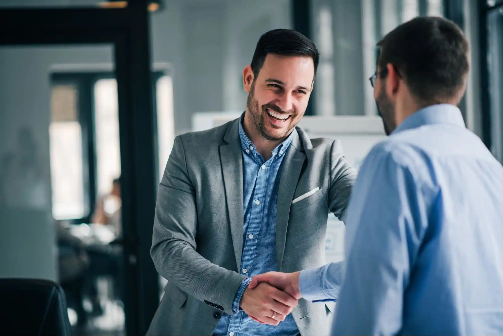 Two men in business attire smiling and shaking hands in a modern office setting, suggesting a successful meeting or agreement.