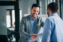 Two men in business attire smiling and shaking hands in a modern office setting, suggesting a successful meeting or agreement.