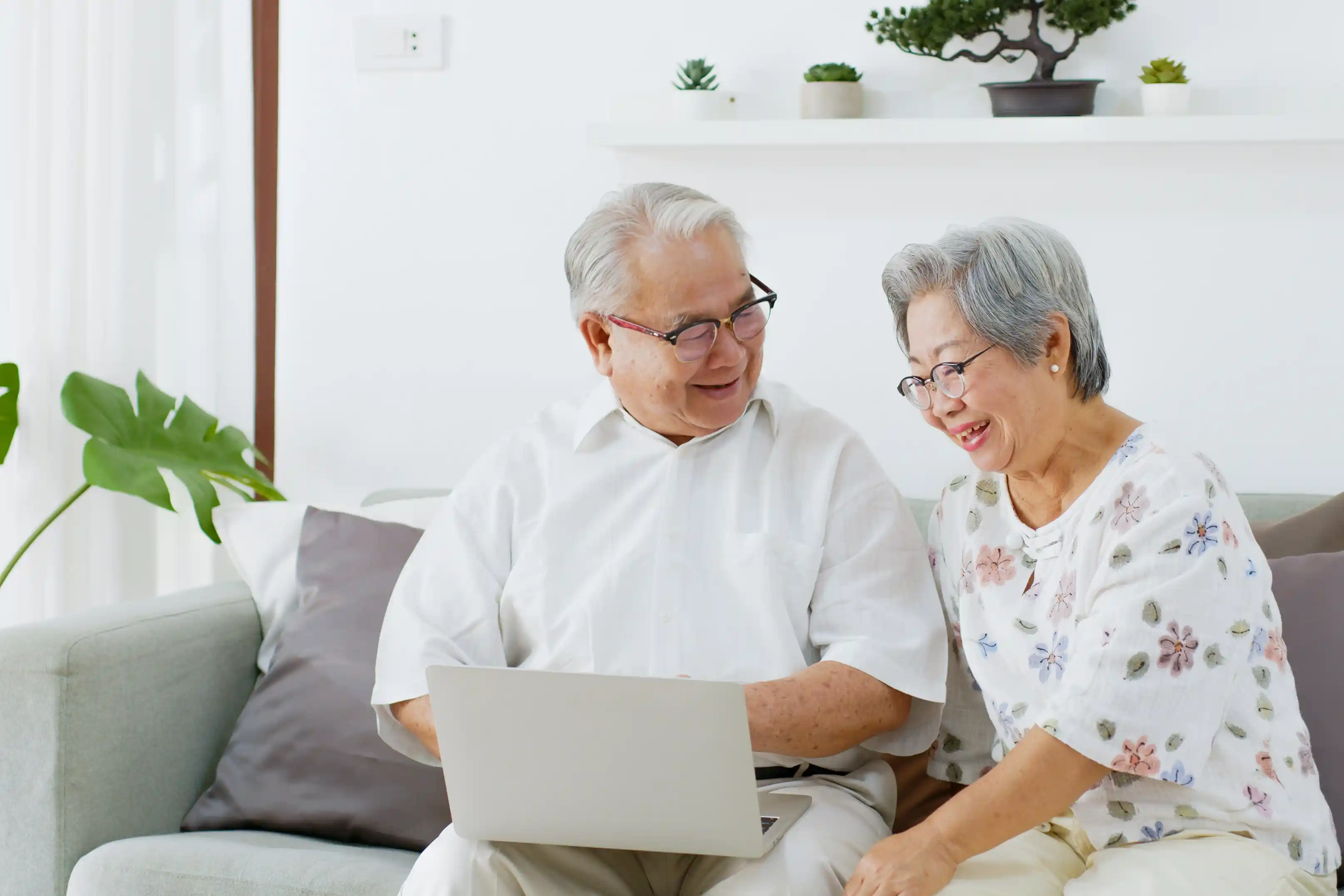 "An elderly couple sitting together on a sofa, smiling and looking at a laptop screen. The man is wearing glasses and a white shirt, while the woman, also wearing glasses, is dressed in a floral-patterned blouse. 