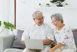 "An elderly couple sitting together on a sofa, smiling and looking at a laptop screen. The man is wearing glasses and a white shirt, while the woman, also wearing glasses, is dressed in a floral-patterned blouse.