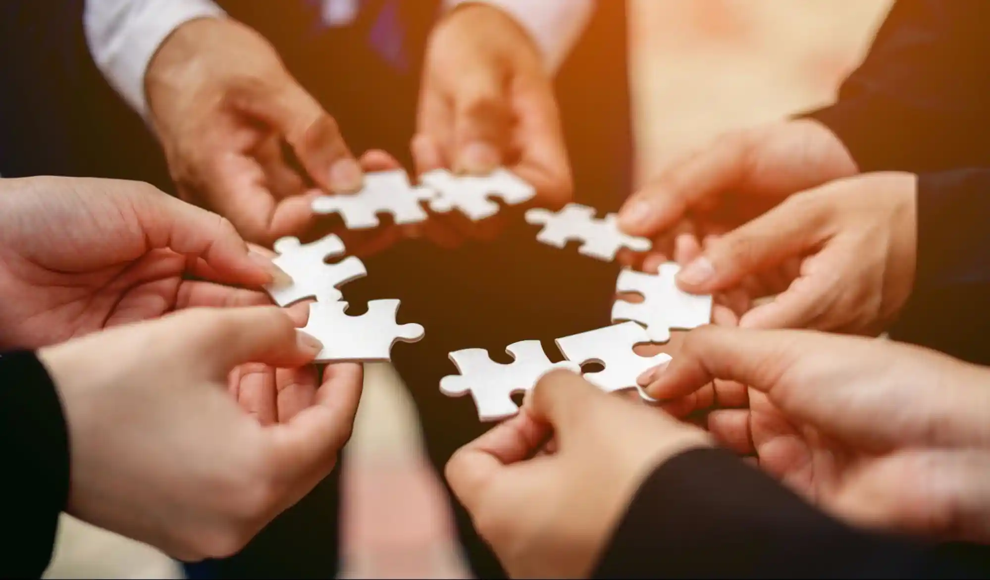A close-up of multiple hands from different individuals, each holding a white puzzle piece, working together to form a complete circle. The image symbolizes teamwork, collaboration, and unity.