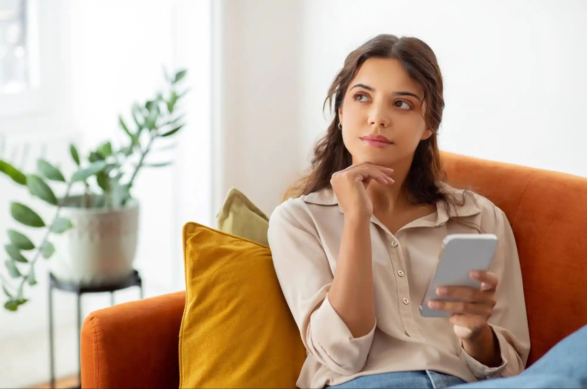 Young woman sitting on an orange couch holding a smartphone, looking thoughtful with her hand resting on her chin, in a bright and cozy living room.