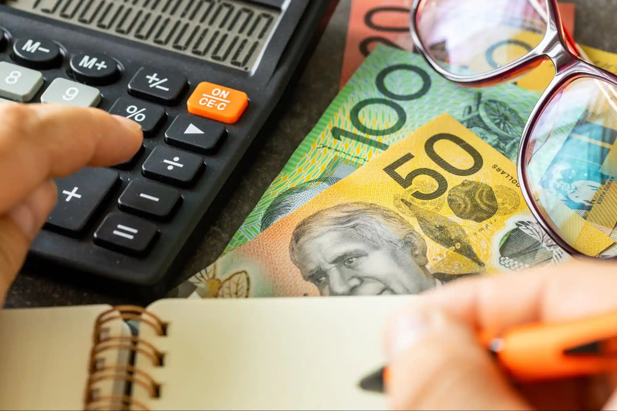 Close-up of a person using a calculator next to banknotes in denominations of 50, 100, and 20, with eyeglasses resting on the money and a hand writing in a spiral notebook.