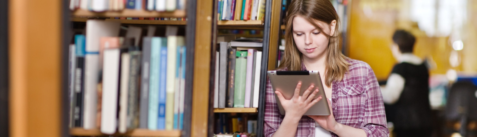 A female test taker standing near a bookshelf  in a library and looking into a tablet.