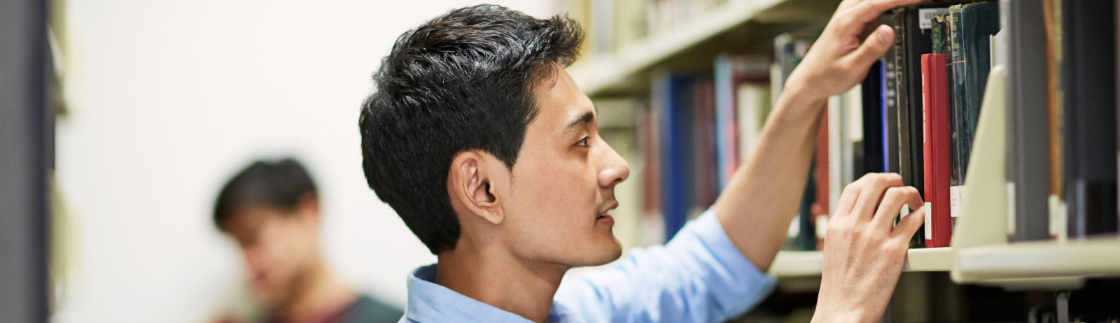 A male IELTS test taker wearing a blue shirt refers to books in a library