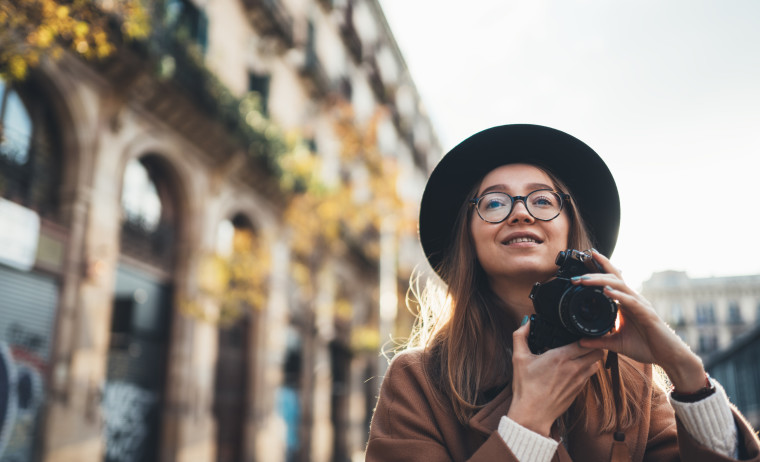Female IELTS test taker wearing a hat, brown coat and spectacles holding a camera in a city street