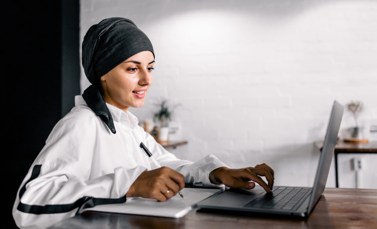 A female IELTS test taker wearing a white shirt and a headscarf practices IELTS on computer.