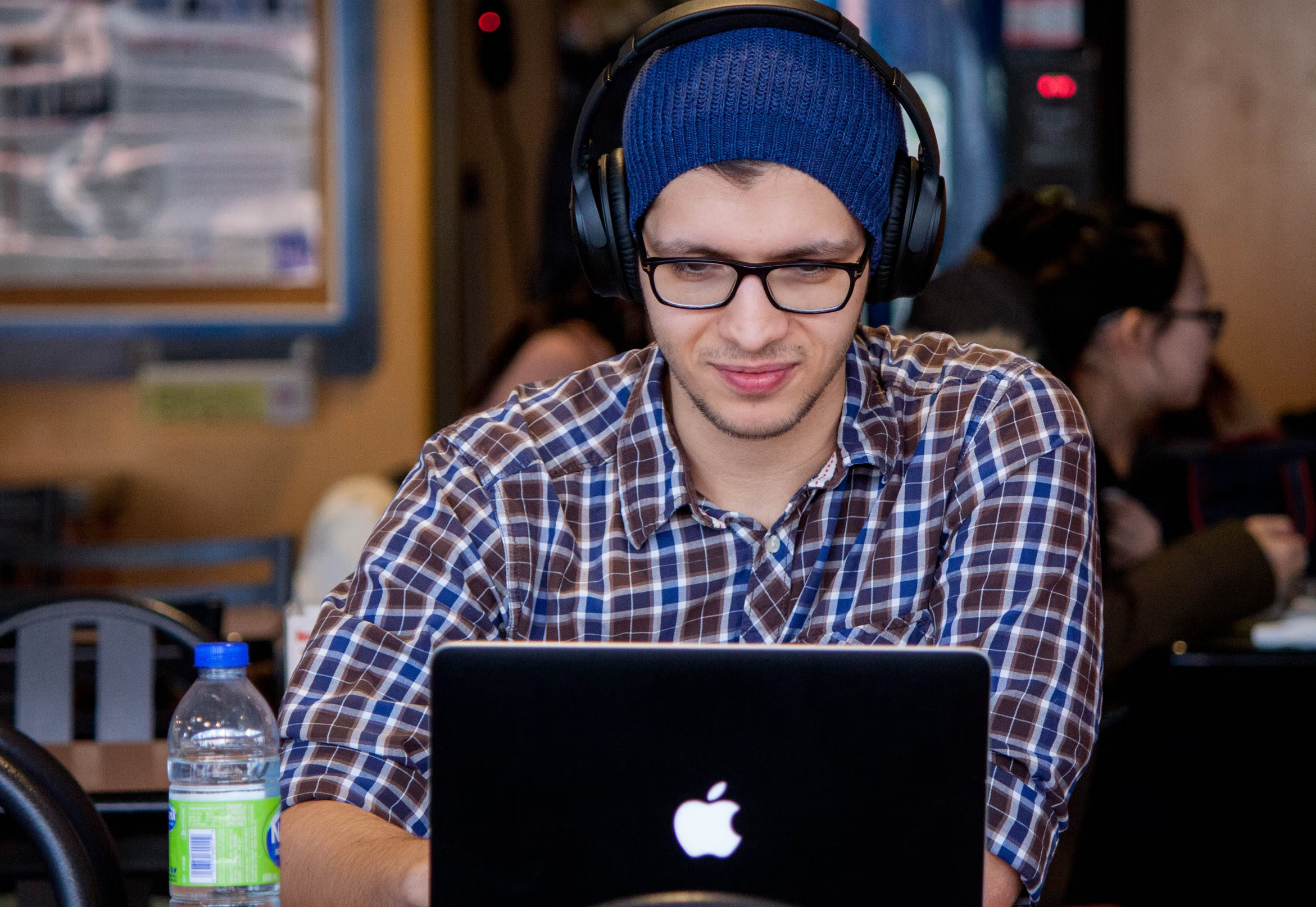 A male IELTS test taker in a cafeteria wearing glasses and headphones and looking into a laptop.