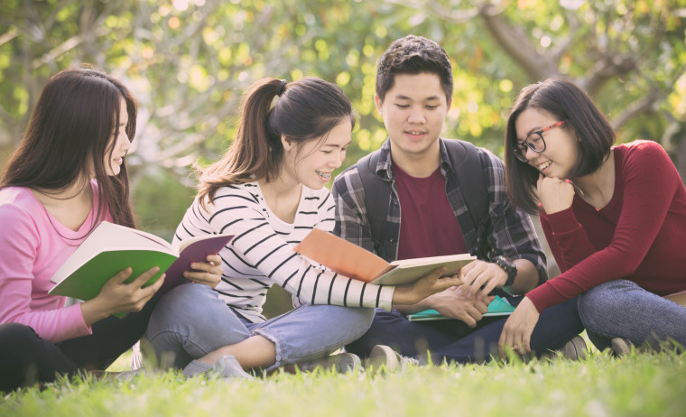 Three female test takers and a male test taker in western outfits siting on grass with IELTS official preparation materials