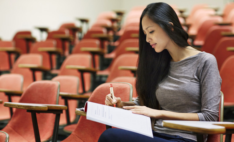 A female test taker in maroon t-shirt sits  in a classroom lecture taking notes for IELTS test preparation.