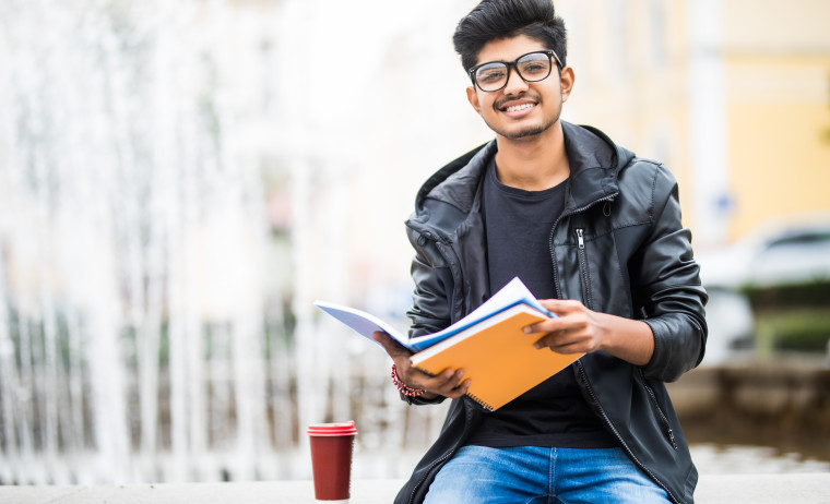 Male IELTS test taker in a black jacket and blue jeans wearing spectacles and reading books, with a coffee cup by his side