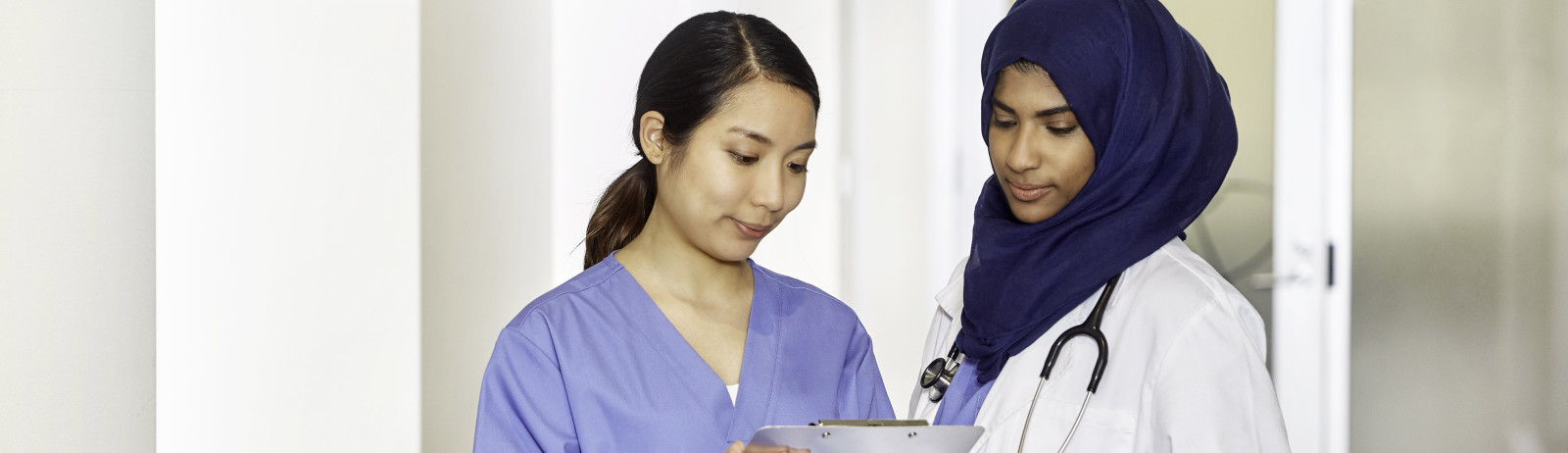 A female nurse and a female doctor wearing a dark blue hijab discuss about a medical record