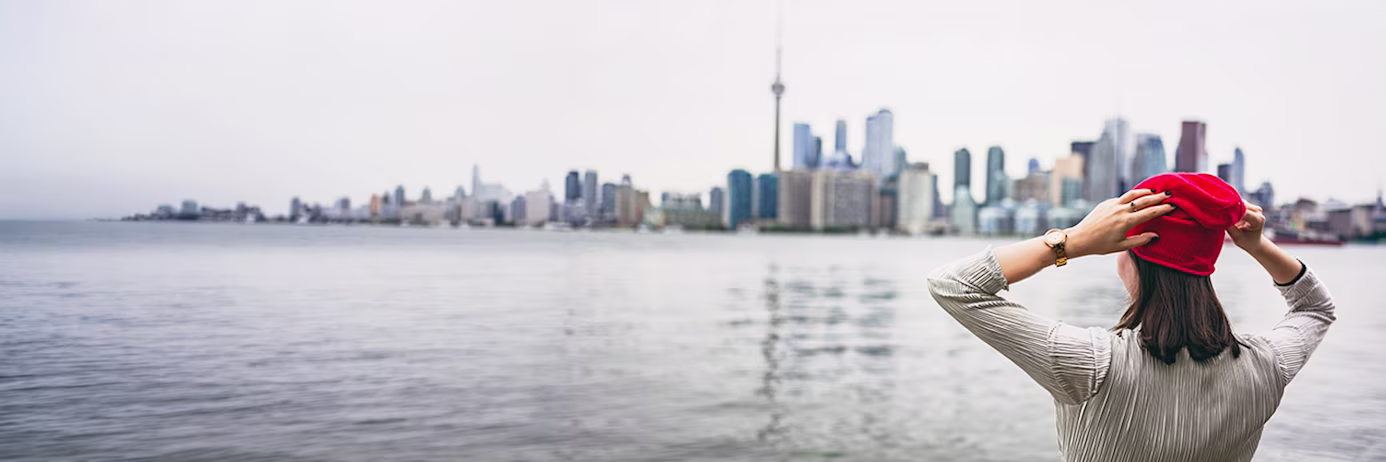 A person looking at the Toronto skyline on a grey day, wearing a red hat