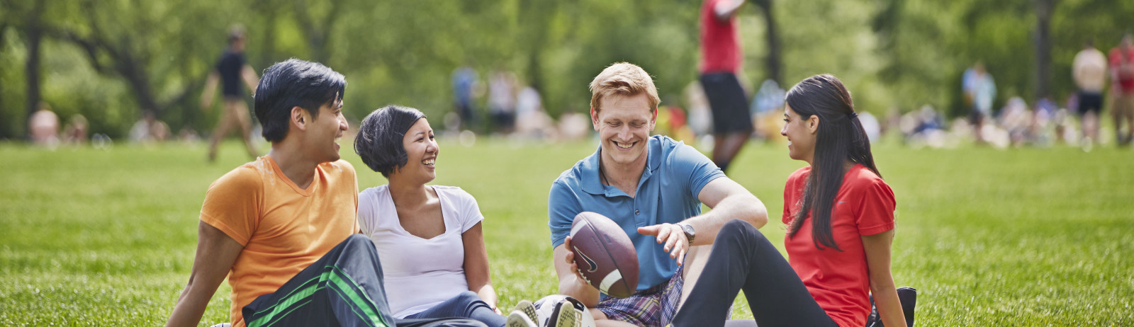 Two female and two male students have a casual talk sitting on the grass with a baseball and a football in a park