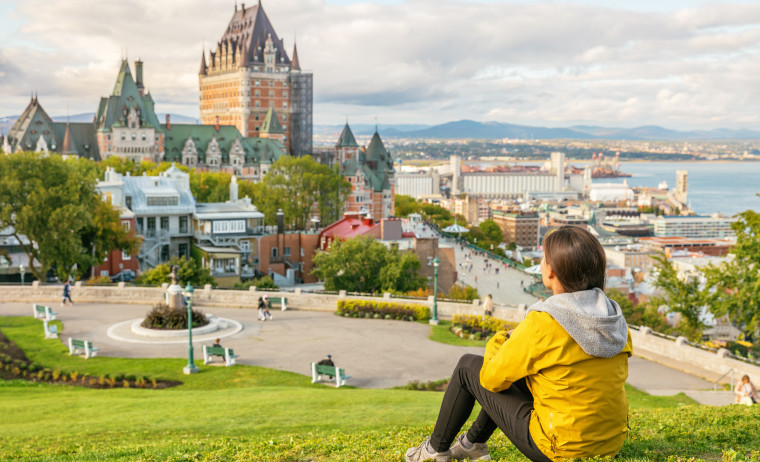 A female IELTS test taker wearing a yellow hoodie sits on grass and enjoys city view 