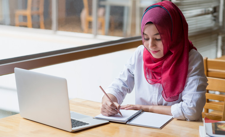 A female IELTS test taker in a pink hijab preparing for the IELTS Writing test in a library