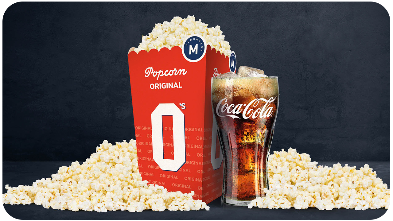 Bilde på et glass med Coca Cola og et rødt beger med popkorn med ODEON Kinos logo på. Mørkeblå bakgrunn og popkorn rundt beger og glass.