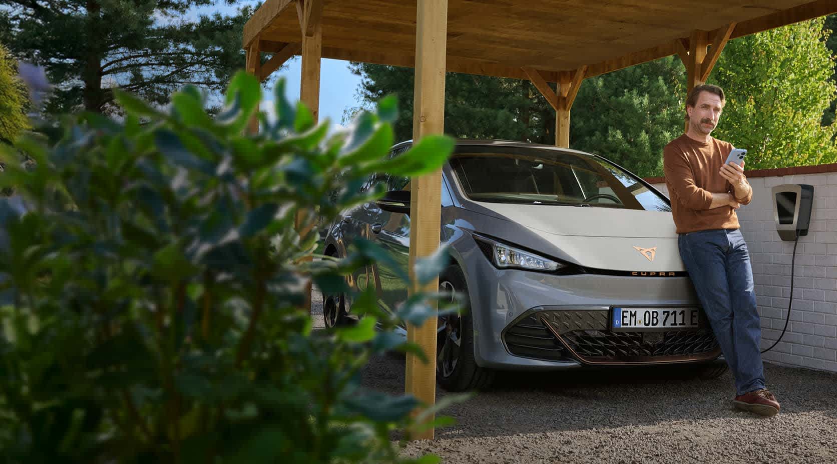 Ein Mann mit Smartphone in der Hand lehnt an der Motorhaube seines E-Autos, das an der Wallbox im Carport lädt.