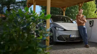 Ein Mann mit Smartphone in der Hand lehnt an der Motorhaube seines E-Autos, das an der Wallbox im Carport lädt.