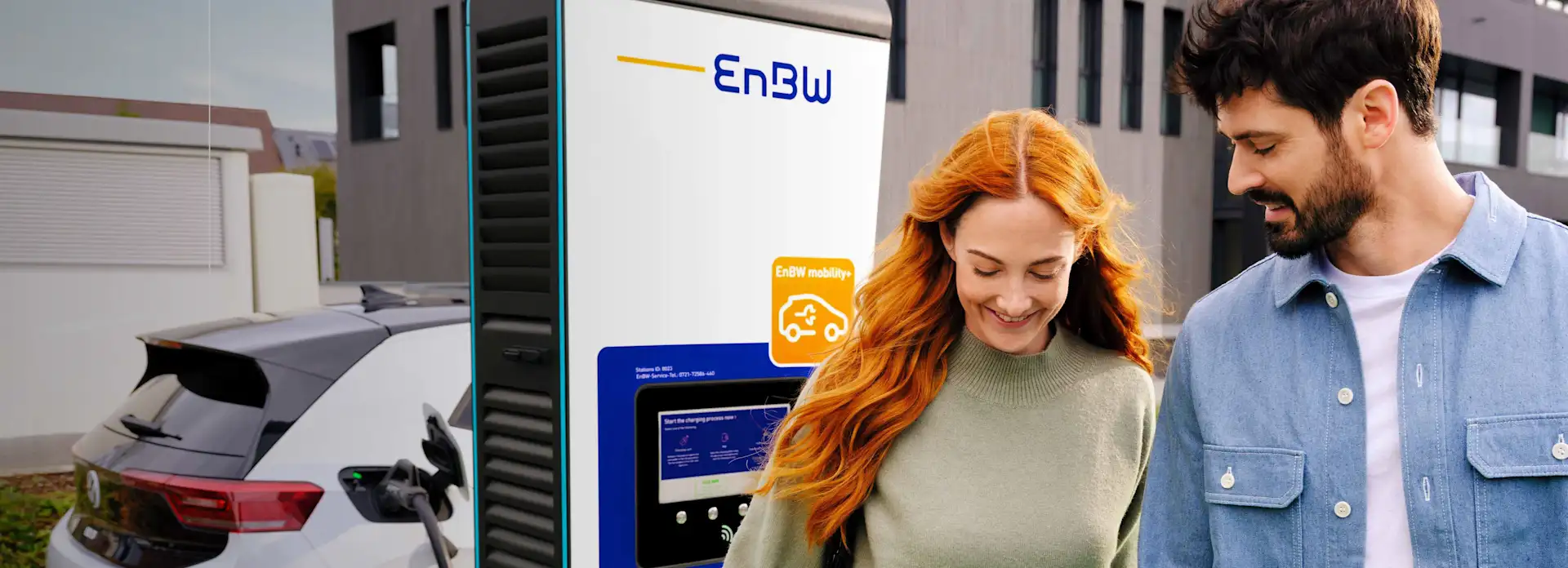 Orangehaarige Frau und Mann vor Ladesäule, Elektroauto lädt im Hintergrund