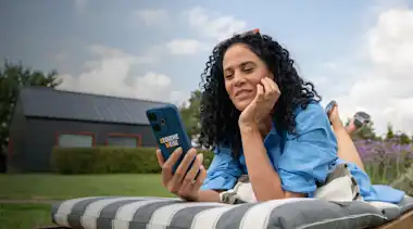 Eine Frau liegt auf einem Liegestuhl im Garten und steuert EnBW Mavi bequem über die zuhause+ App auf ihrem Smartphone.