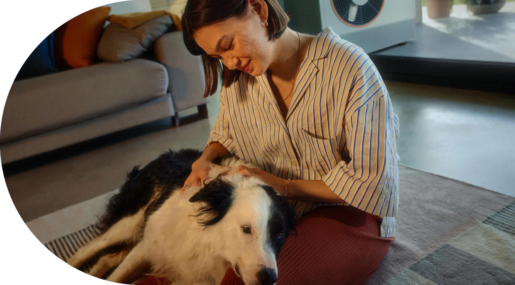 Junge Frau liegt mit ihrem Hund auf einem Teppich im Wohnzimmer ihres Hauses. Im Hintergrund auf der Terrasse ist eine Wärmepumpe zu sehen.