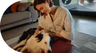 Junge Frau liegt mit ihrem Hund auf einem Teppich im Wohnzimmer ihres Hauses. Im Hintergrund auf der Terrasse ist eine Wärmepumpe  zu sehen.