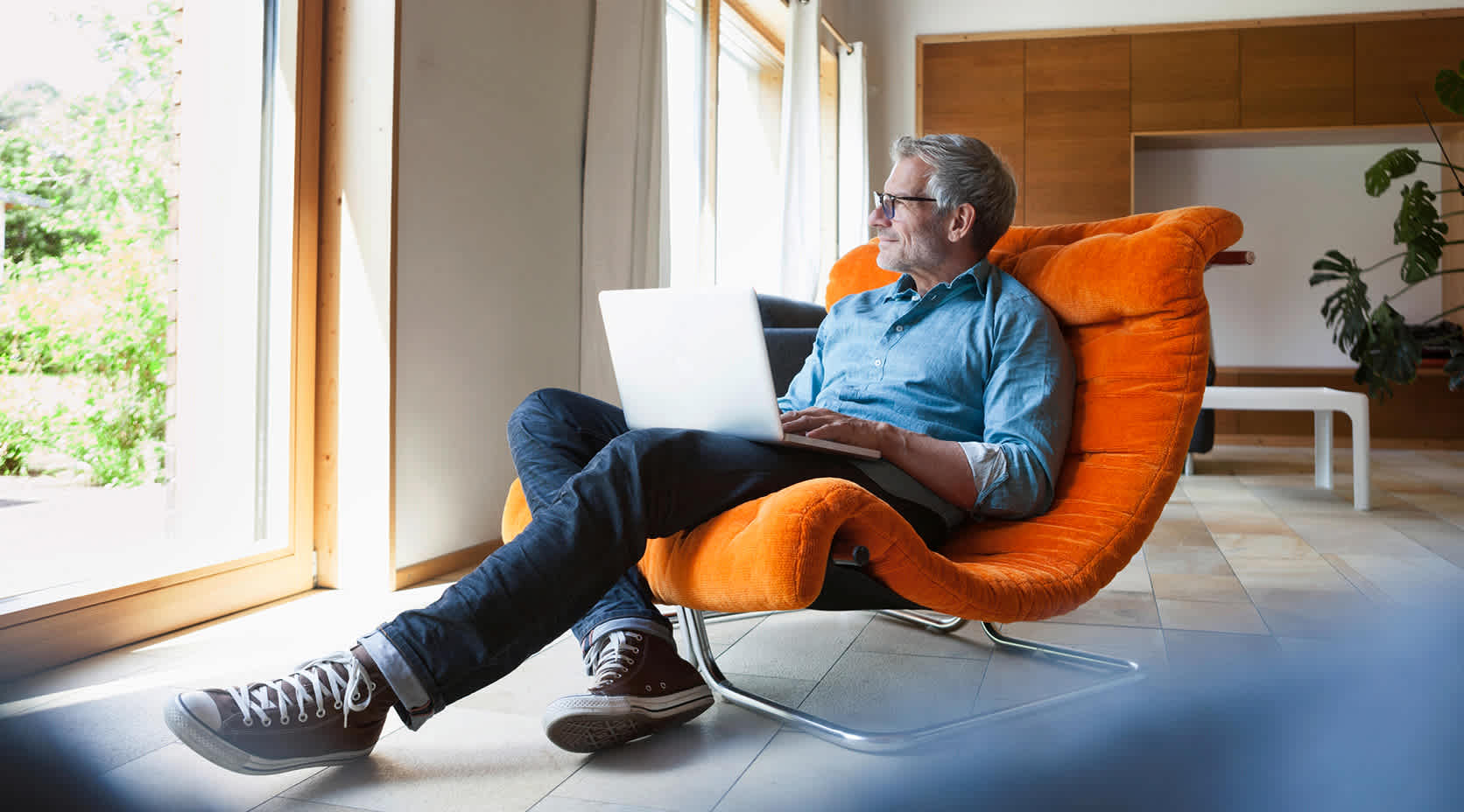 Älterer, zufriedener Mann in seinem Zuhause auf einem orangenem Sessel mit Laptop.