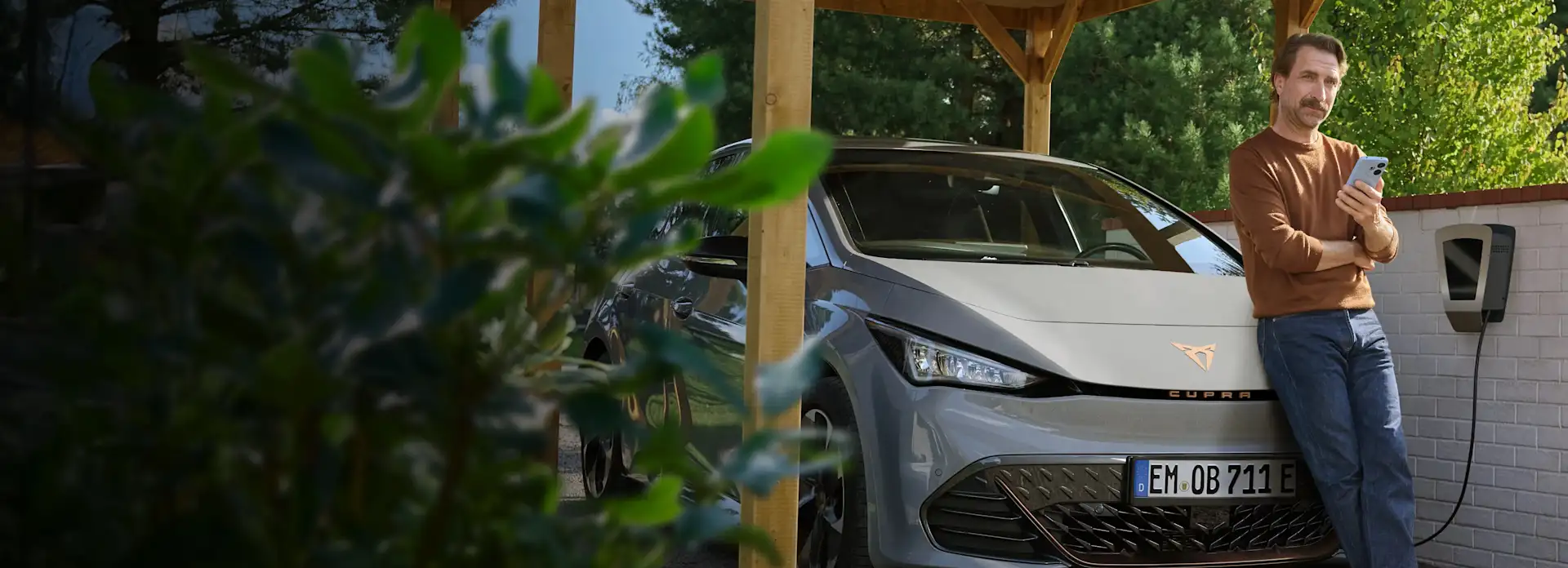 Ein Mann mit Smartphone in der Hand lehnt an der Motorhaube seines E-Autos, das an der Wallbox im Carport lädt.