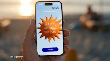 Eine Hand, die ein Smartphone festhält. Im Hintergrund befindet sich eine Strandkulisse mit Meer, Sand und Liegestühlen. Der Screen auf dem Smartphone zeigt einen Luftballon in Form einer Sonne mit der Aufschrift "5 Cent weniger pro kWh".