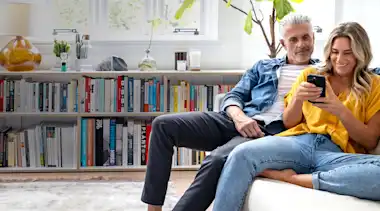 Paar im mittleren Alter sitzt auf der Couch und schaut auf ein Smartphone.