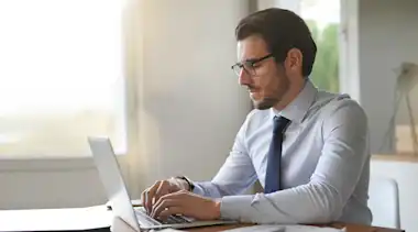 Dunkelhaariger Mann mit Brille, Hemd und Krawatte am Laptop am Schreibtisch