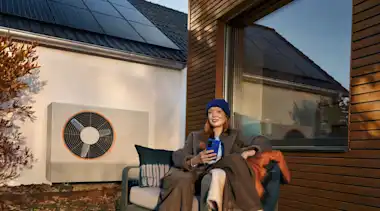 KI-generiert: Eine Frau sitzt im Garten auf einer Bank mit vielen Decken und Kissen. Im Hintergrund sind eine Holzhausfassade und eine Wärmepumpe zu sehen.