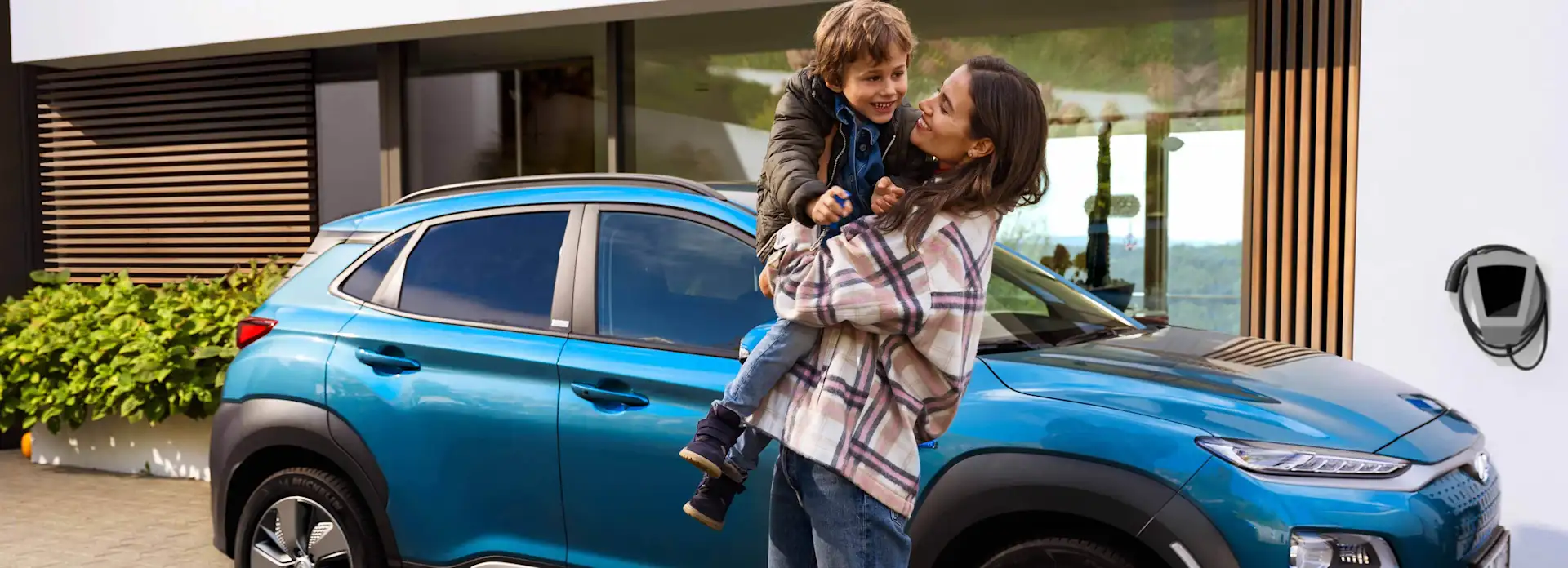 Mutter hat Sohn auf dem Arm vor blauem Auto und Haus mit Wallbox