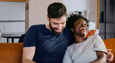 Ein junges Pärchen sitzt lachend auf der Couch und schaut auf ein Smartphone.