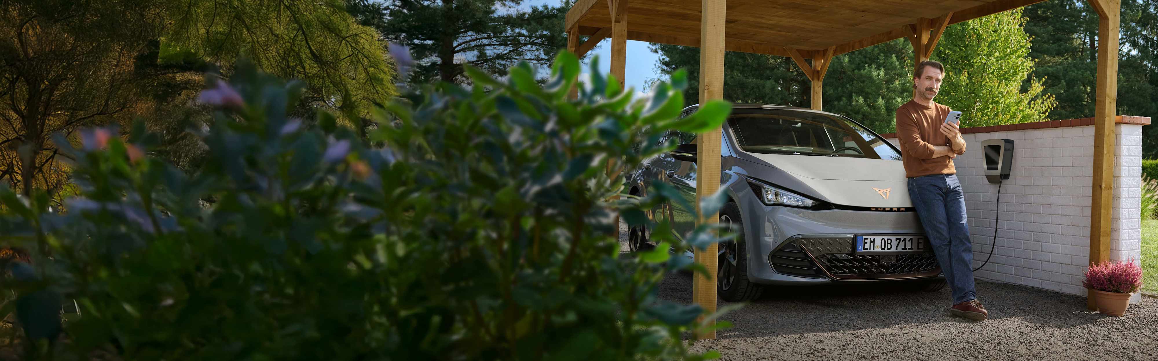 Ein Mann mit Smartphone in der Hand lehnt an der Motorhaube seines E-Autos, das an der Wallbox im Carport lädt.