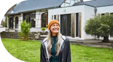 Eine ältere Frau mit langen Haaren steht lächelnd in einem Garten mit Altbau im Hintergrund.