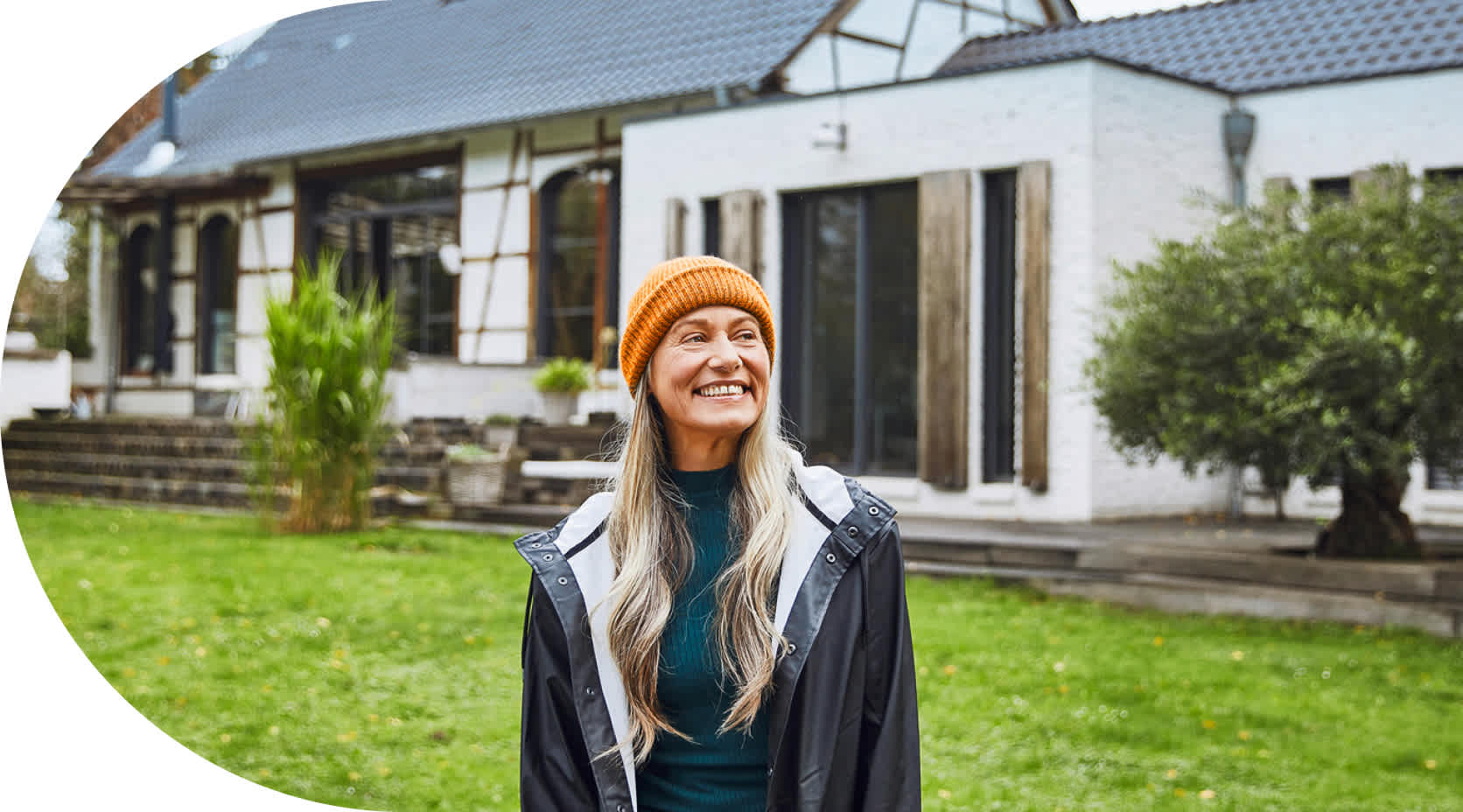 Eine ältere Frau mit langen Haaren steht lächelnd in einem Garten mit Altbau im Hintergrund.