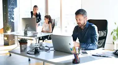 Mann sitzt am Schreibtisch und arbeitet am Laptop. Im Hintergrund unterhalten sich zwei Frauen am Schreibtisch.