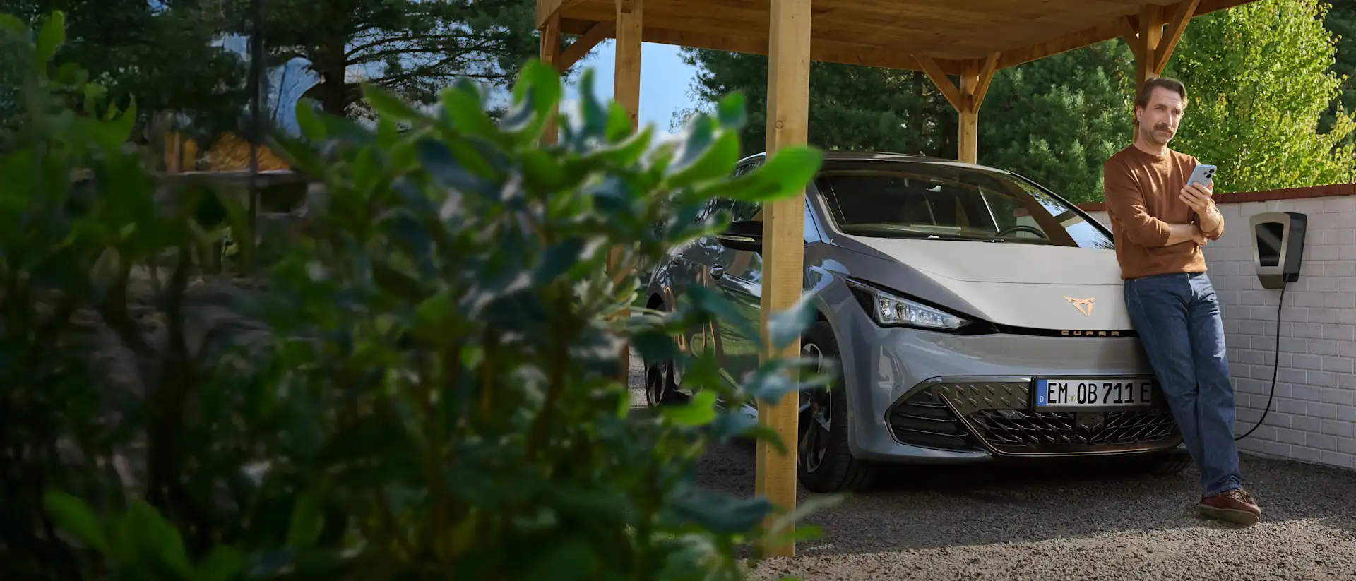 Ein Mann mit Smartphone in der Hand lehnt an der Motorhaube seines E-Autos, das an der Wallbox im Carport lädt.