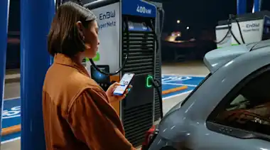 Eine junge Frau lädt an einer EnBW Ladesäule und schaut auf ihr Smartphone.