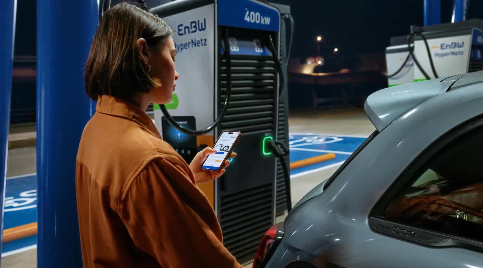 Eine junge Frau lädt an einer EnBW Ladesäule und schaut auf ihr Smartphone.