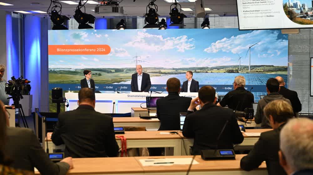 Zuschauende bei einer Pressekonferenz mit 3 präsentierenden Personen vor einer großen Bildleinwand