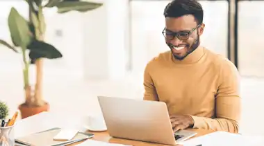 Junger Mann mit Brille lacht Laptop am Schreibtisch an mit Pflanze im Hintergrund